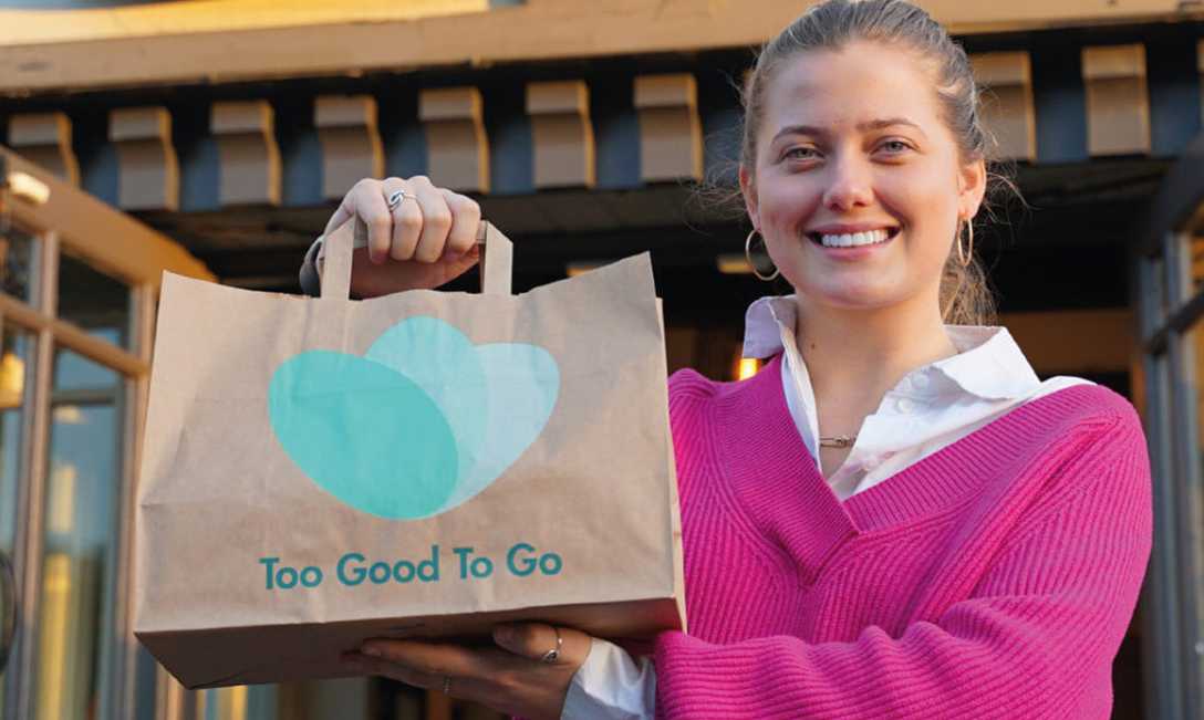 A smiling woman holds up a paper "Too Good To Go" bag outside a restaurant.