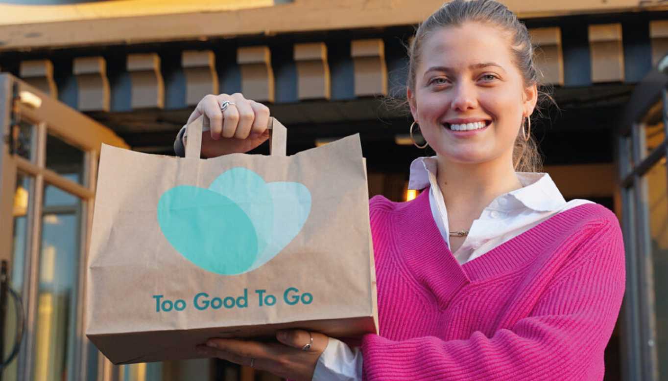 A team member in a bright pink jumper holds a Too Good To Go takeaway bag, representing efforts to avoid food waste.