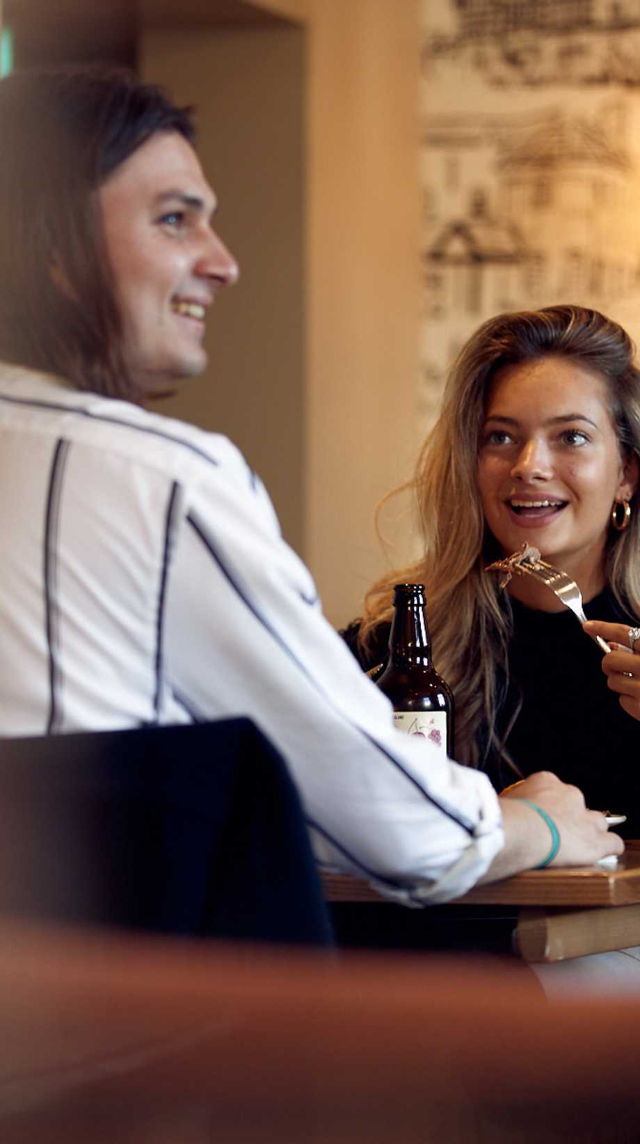 A woman is lifting her fork to her mouth, smiling, while looking at her partner, who is smiling at something off-camera.