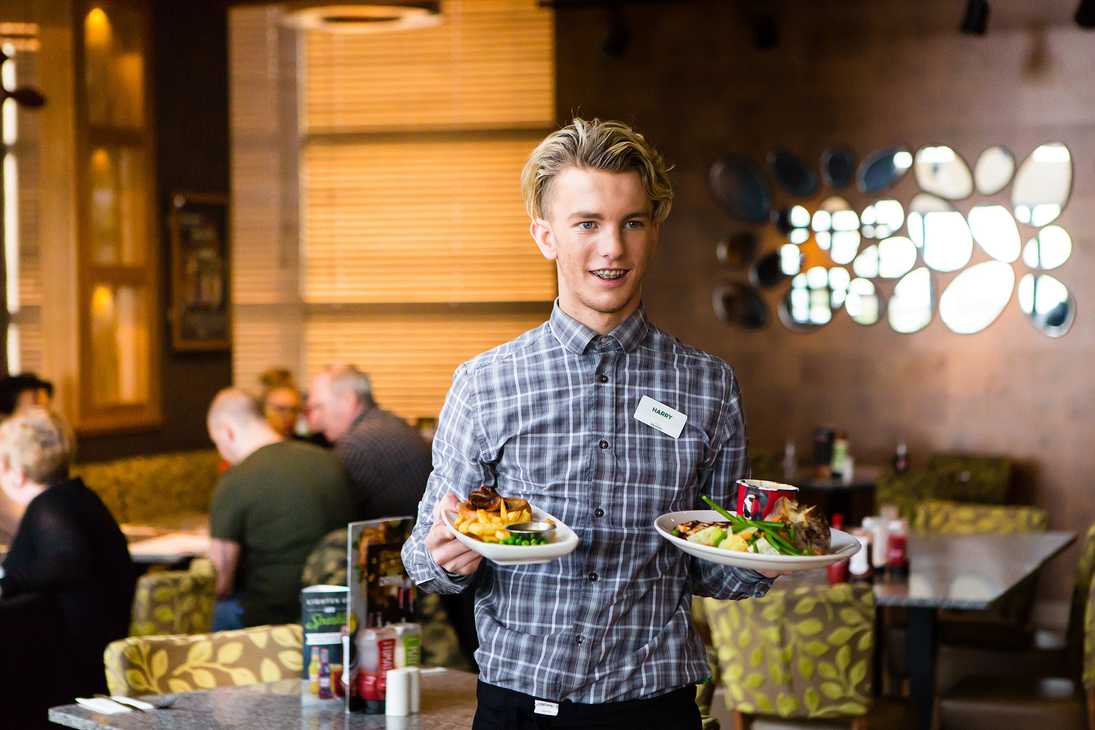 A team member carries two meals to waiting guests. Behind him, seated guests are browsing a menu.