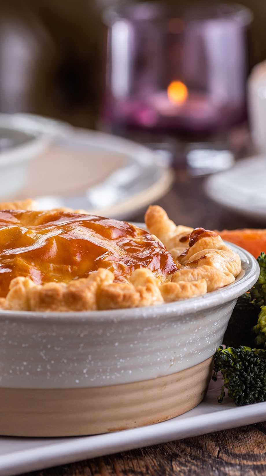 A cosy pie and colourful carrots and brocolli sit in the foreground. In the background, coffee and dessert are waiting.