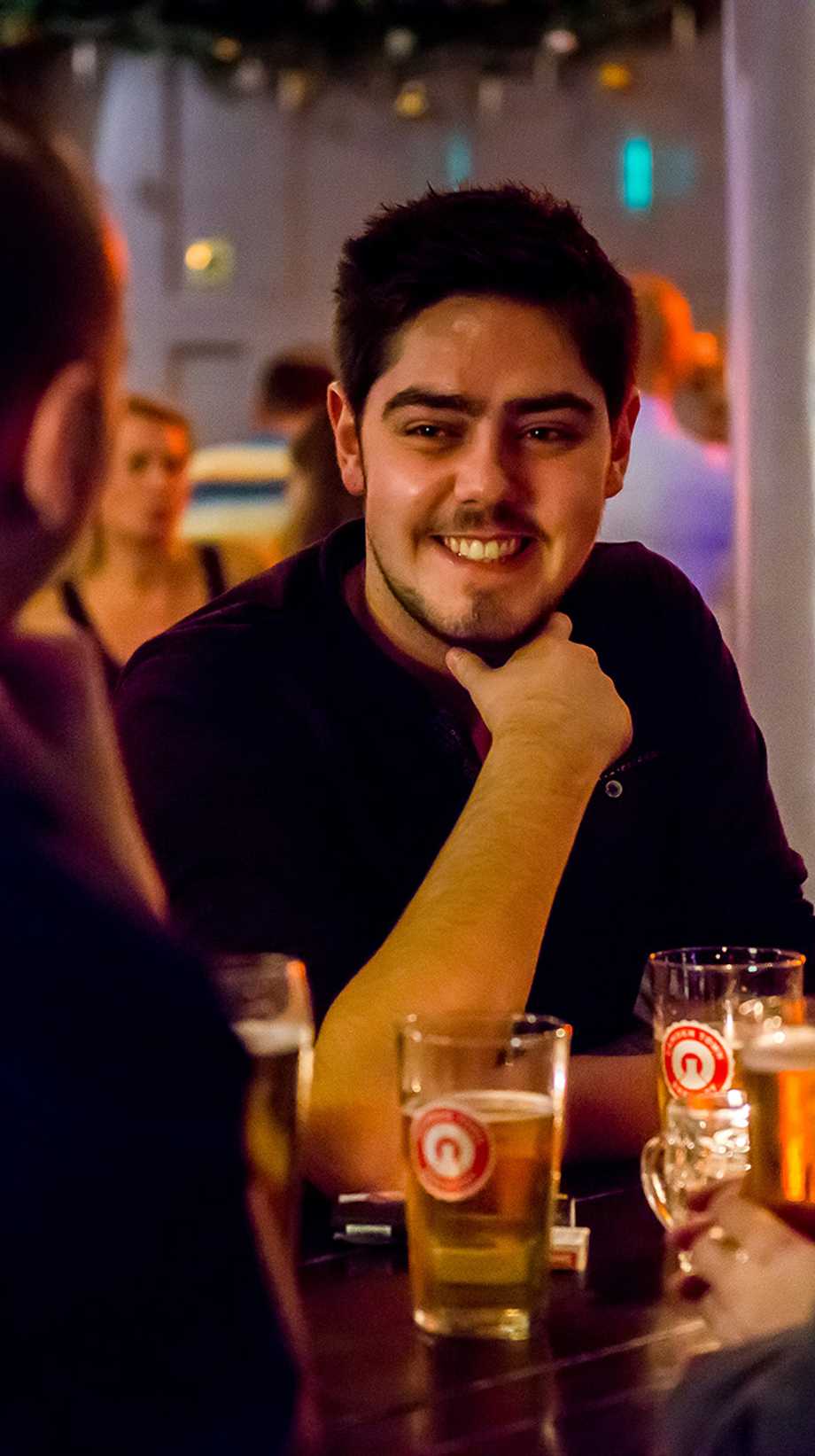 Three friends are chatting over pints. In the background, there are more guests and a glowing neon sign.