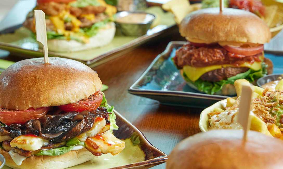 A close-up shot of burgers of different types. In the foreground, a burger is filled with meat, mushrooms, tomatoes and more. Around it, three other burgers are plated.