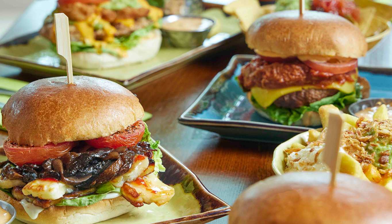 A close-up shot of burgers of different types. In the foreground, a burger is filled with meat, mushrooms, tomatoes and more. Around it, three other burgers are plated.