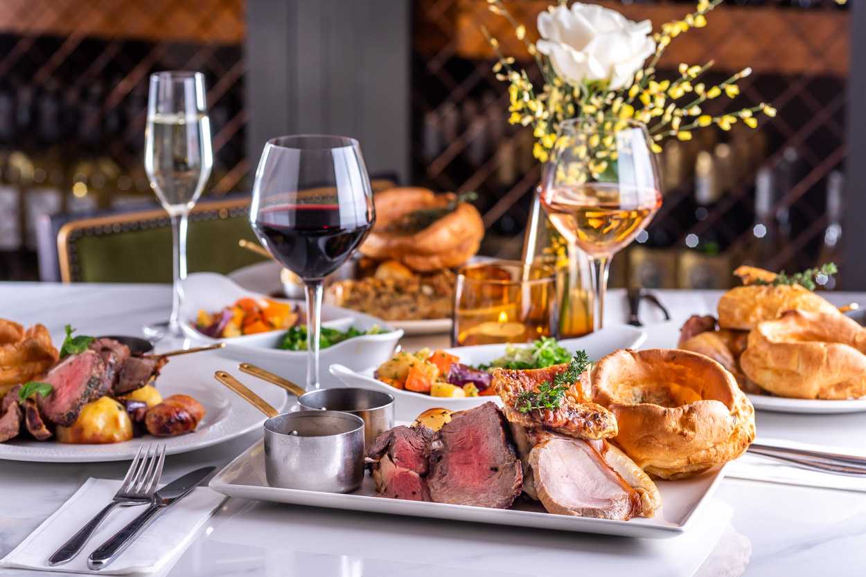 Four roast dinners have been placed on a table, alongside a selection of drinks and white and yellow flowers in the centre.